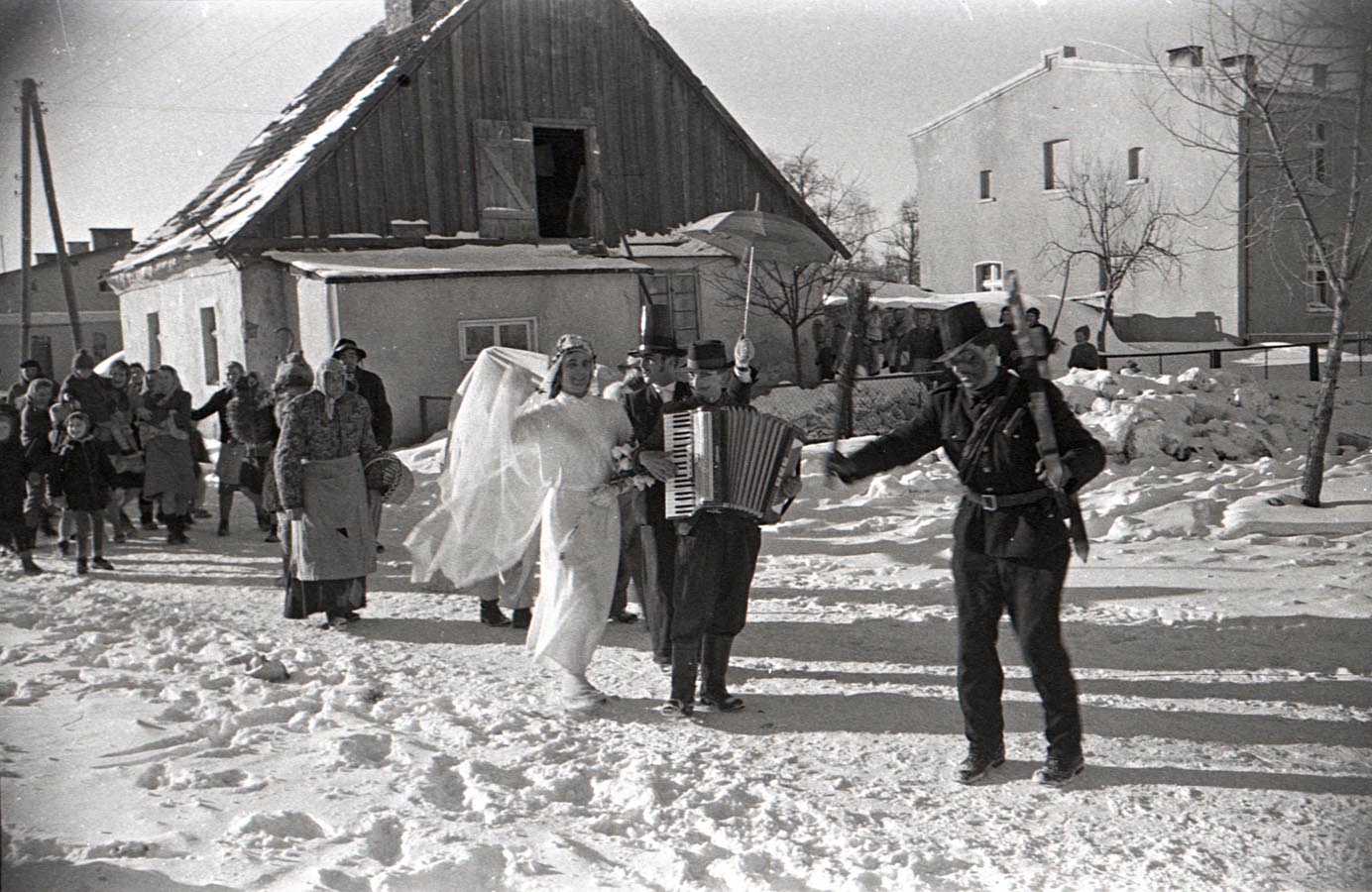 Fotografia czarno-biała przedstawiająca korowód przebierańców idących drogą. Na ich czele widoczny kominiarz i dwie osoby przebrane za parę młodą, mężczyzna gra na akordeonie. Dookoła leży śnieg, w tle widoczne budynki mieszkalne.