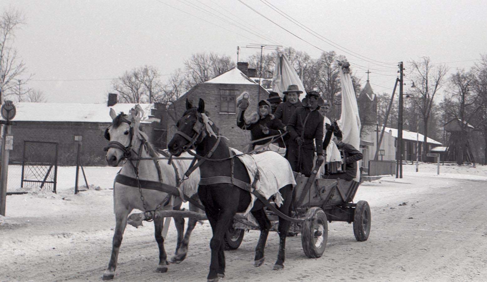 Fotografia czarno-biała. Zima, grupa przebranych osób jedzie drewnianym wozem zaprzęgniętym w dwa konie, najprawdopodobniej siwka i karego. W tle widać zabudowania mieszkalne, lampy uliczne i wieżę kościoła.