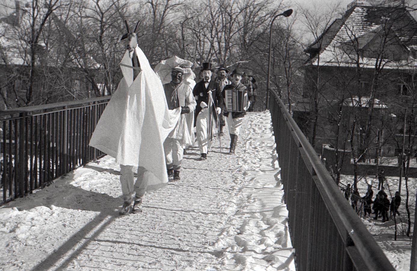 Fotografia czarno-biała przedstawiająca grupę przebranych osób idących ośnieżonym mostkiem. Na czele korowodu widoczny kozioł, w tle widać osobę grającą na akordeonie. Na dalszych planach dostrzec można także budynki, przechodniów i drzewa.