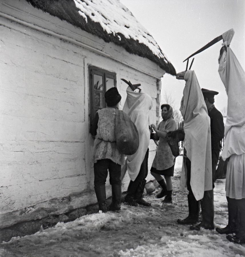 Fotografia czarno biała. Grupa przebranych osób stoi pod oknem tradycyjnego domu.