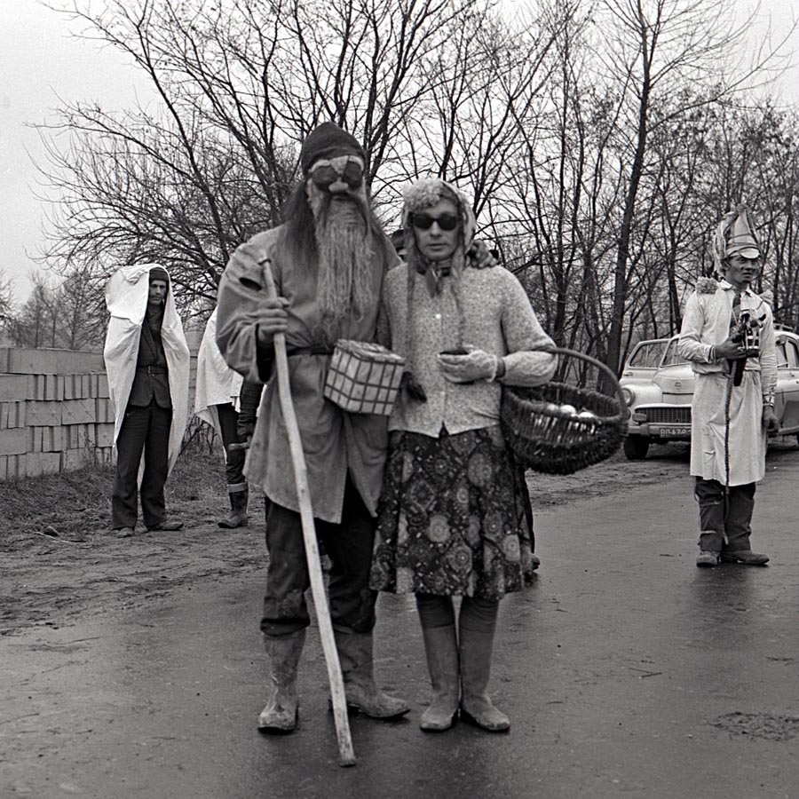 Fotografia czarno- biała, na której widać pozujące do zdjęcia dwie przebrane osoby. W tle widać murowany płot, drogę, samochód i innych przebierańców.