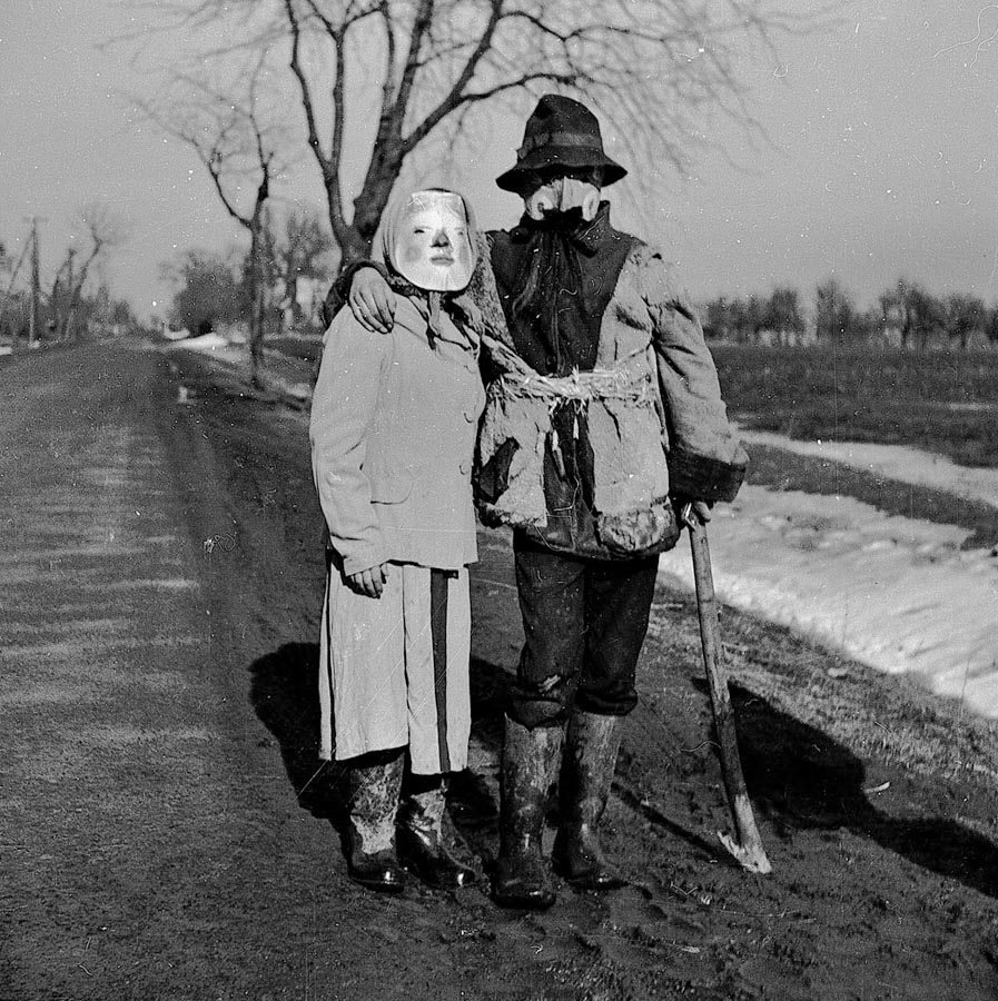 Fotografia czarno-biała. Dwójka przebierańców pozuje do zdjęcia na wiejskiej drodze. Osoba po lewej ubrana w spódnicę i marynarkę, a na głowie ma maskę i chustę. Osoba po prawej ubrana jest natomiast w kożuch, a na jej głowie znajduje się maska i kapelusz. W tle widoczne drzewa i pola uprawne.
