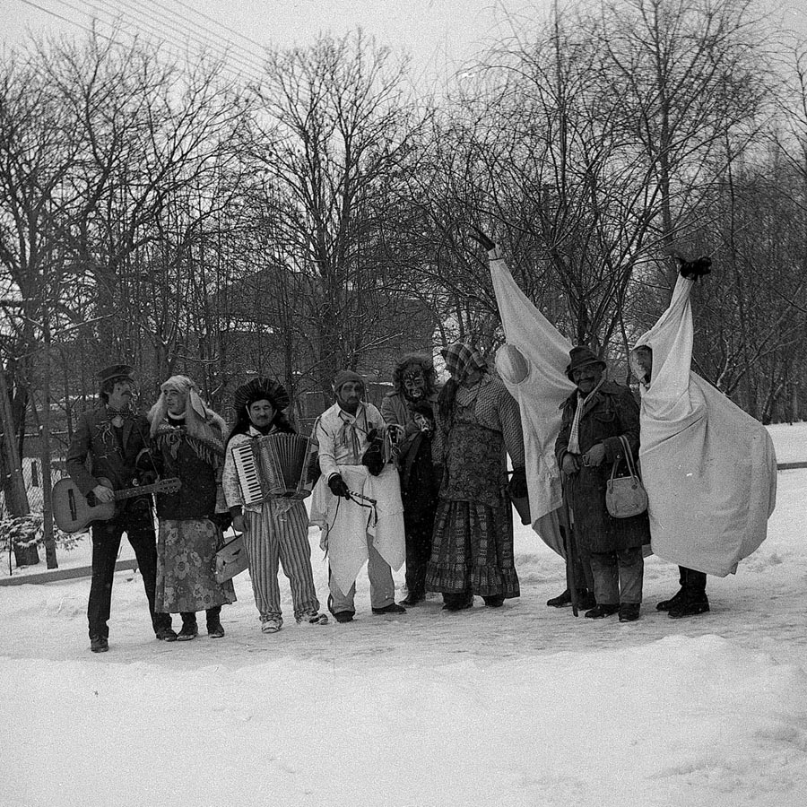 Fotografia czarno-biała. Widok na grupę przebranych osób, najprawdopodobniej zimową porą. Wśród nich znajdują się dwa kozły, diabeł i trójka osób grających na instrumentach. W tle widoczne drzewa i zabudowania.