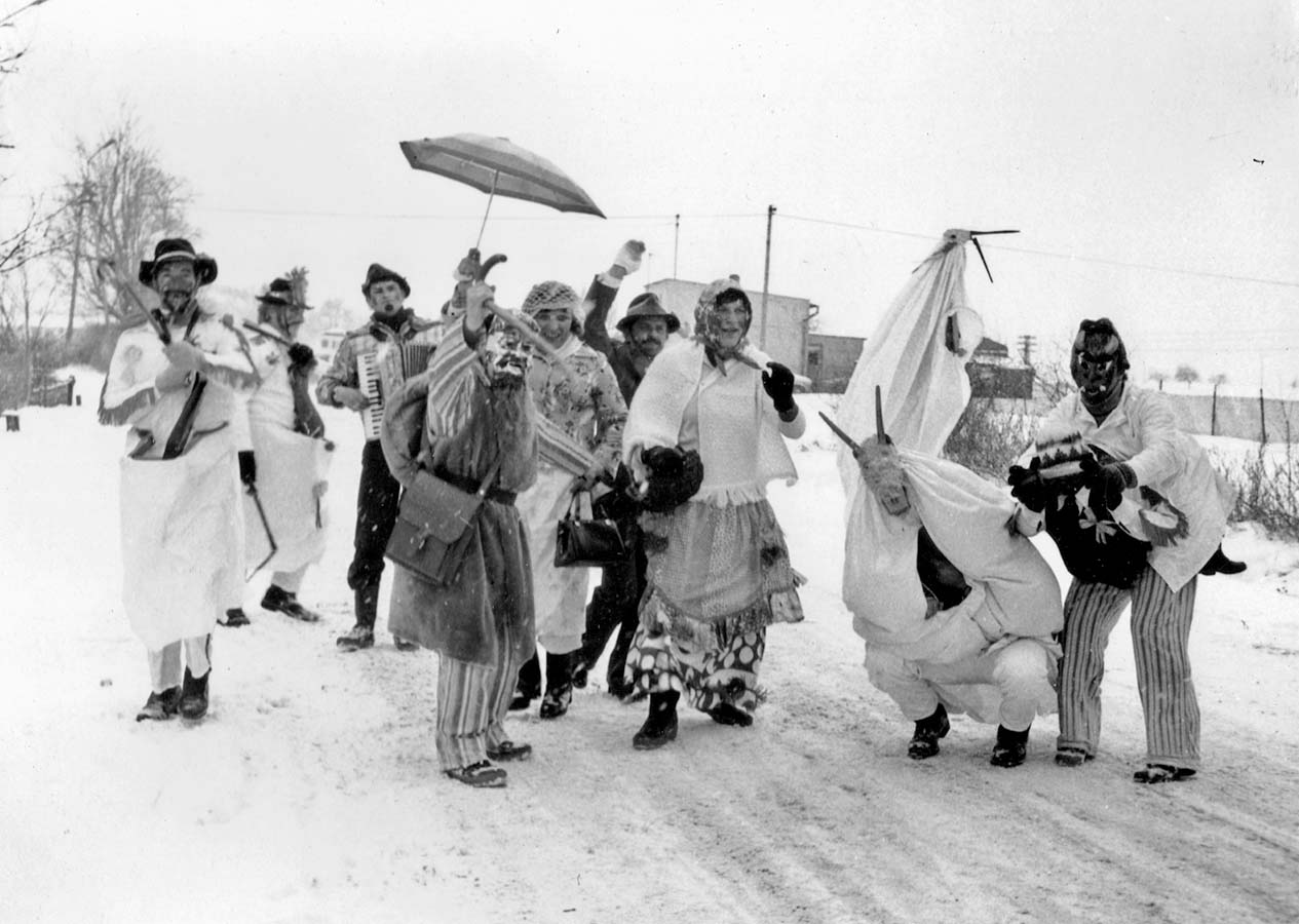 Fotografia czarno-biała przedstawiająca grupę przebranych osób podczas zabawy. Przebierańcy znajdują się na drodze, dookoła leży śnieg a w tle widoczne są zabudowania.
