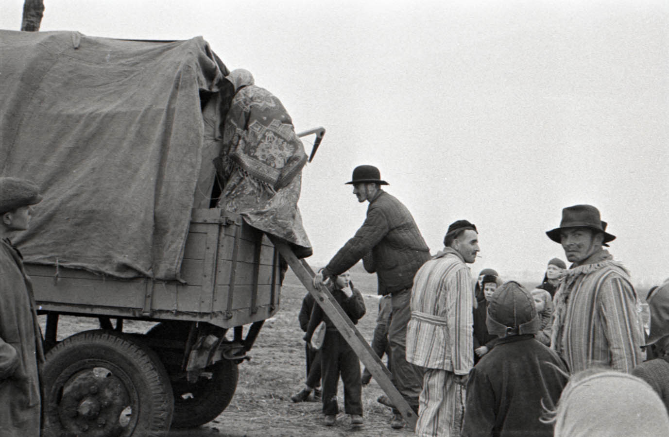 Fotografia czarno-biała przedstawiająca wsiadających do drewnianego wozu przebierańców. W tle widoczne pola uprawne.